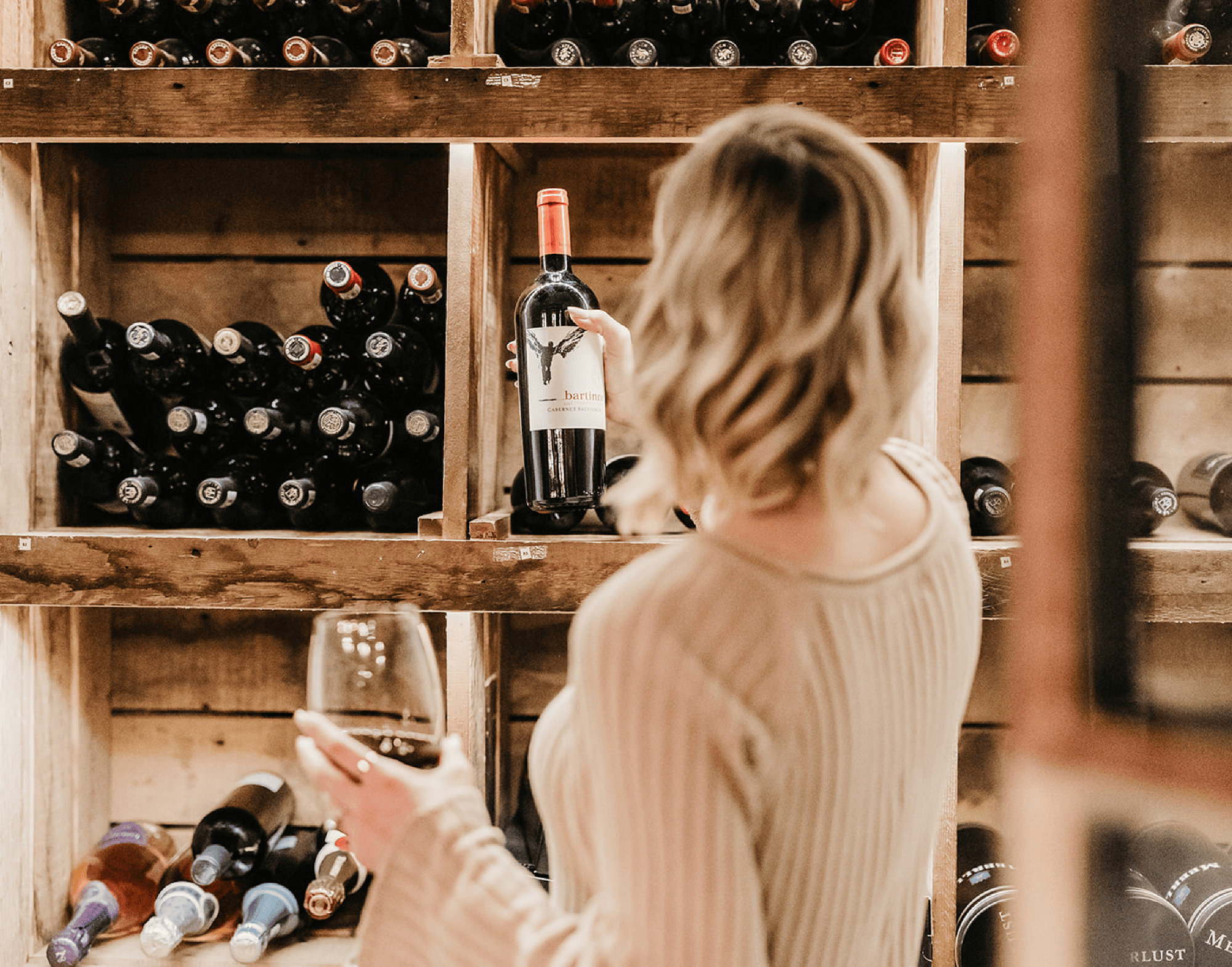 Woman selecting a bottle of red wine from a wooden wine rack; Honesty Bar