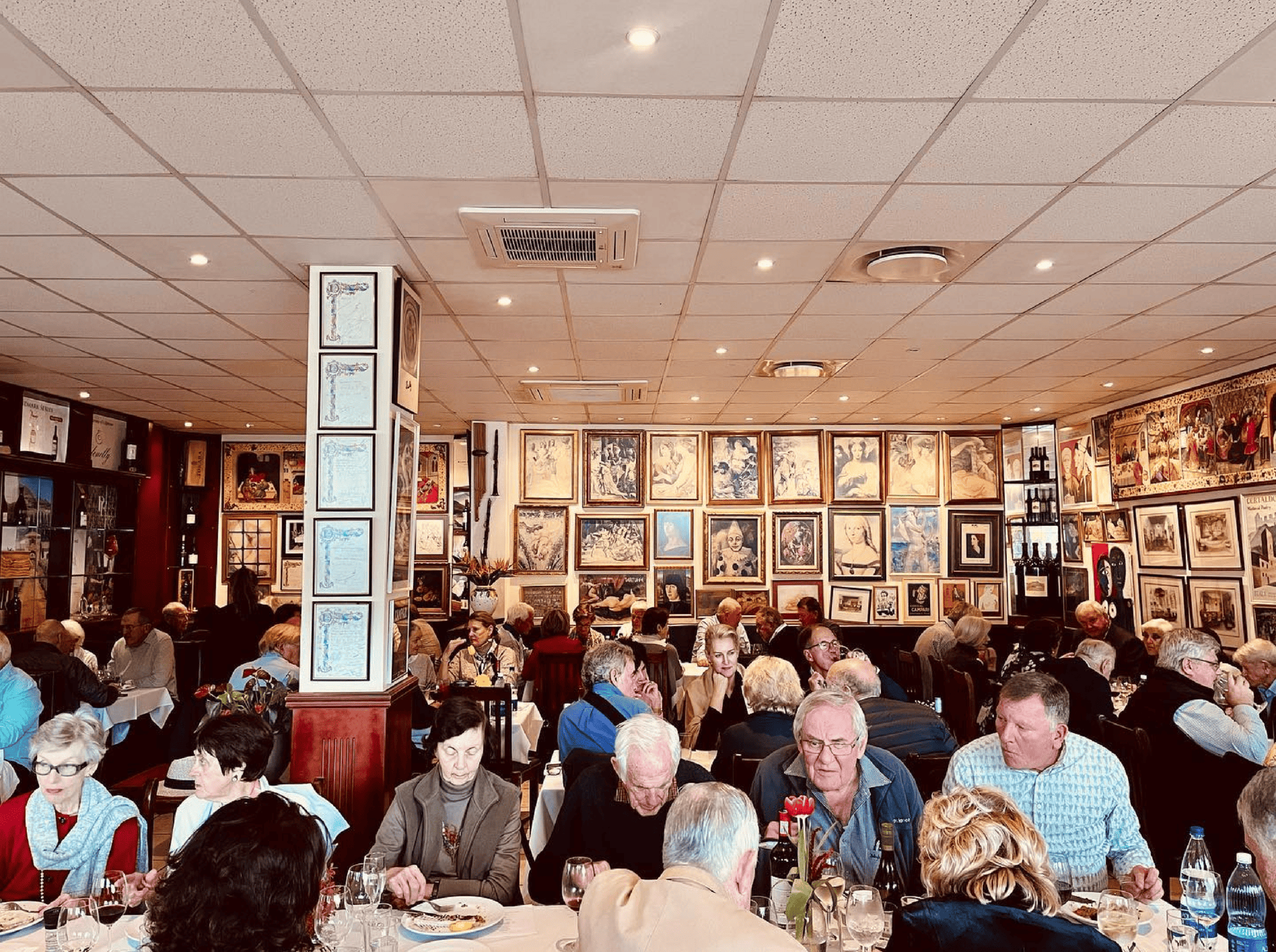 People dining at tables in a restaurant with framed art on the walls, seen as you explore the Cape Winelands