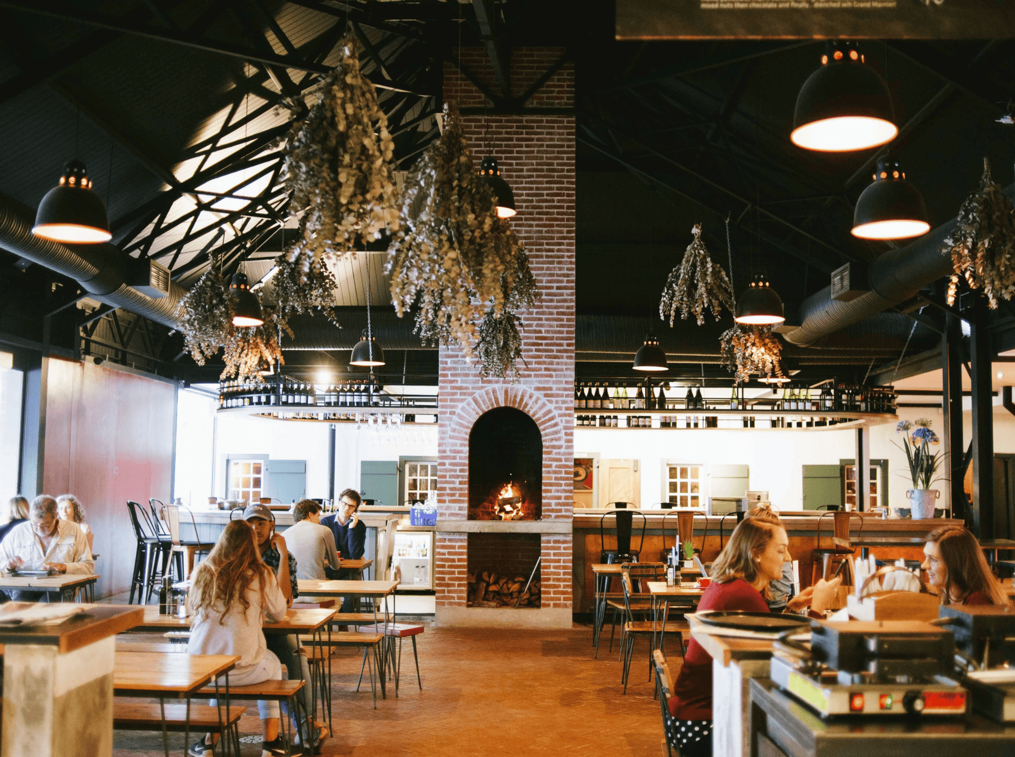 People dining in a rustic restaurant with a large fireplace and hanging dried herbs.